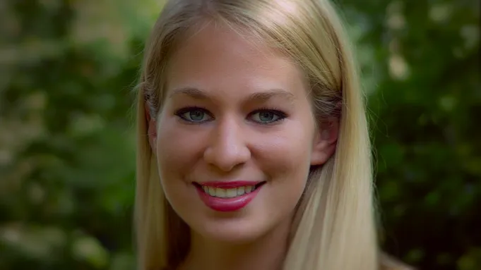 "Smiling young woman with long blonde hair outdoors, green blurred foliage in the background, natural lighting, close-up portrait."